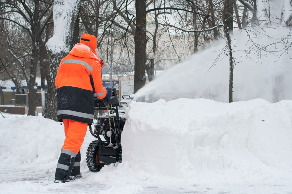Gemeindearbeiter in oranger Schutzkleidung mit Schneefräse bearbeitet verschneite Parklandschaft